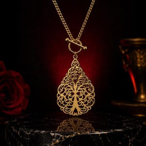 Peut inclure: Un collier doré composé d'un pendentif en forme de goutte ajourée avec un motif complexe d'arbre en filigrane. Le pendentif est suspendu à une chaîne dorée assortie, fixée par un fermoir à barre horizontale et anneau.