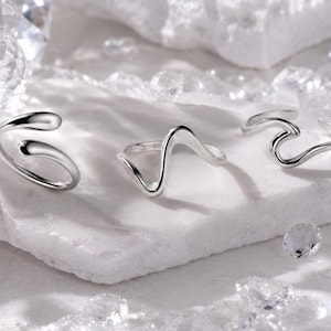 Peut inclure: Trois bagues argentées aux designs fluides et uniques sont présentées sur une surface blanche. Les bagues ont une esthétique moderne et minimaliste, avec des lignes lisses et courbes. Le fond présente des cristaux étincelants, rehaussant l'élégance des bijoux.