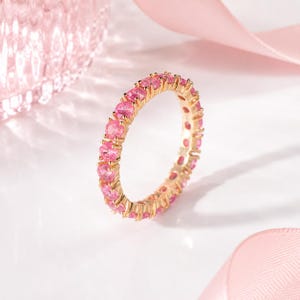 Peut inclure: Une bague en or ornée d'une rangée continue de pierres précieuses roses de forme ovale. La bague est posée sur un fond rose clair avec un ruban et un objet décoratif flou, créant une esthétique délicate et féminine.