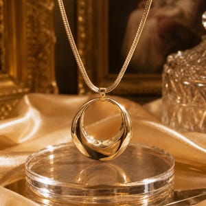 Peut inclure: Un collier doré doté d'un pendentif circulaire poli avec un centre évidé. Le pendentif est suspendu à une chaîne en maille serpent plate et flexible. La surface métallique est lisse et réfléchissante, captant la lumière pour créer des reflets.