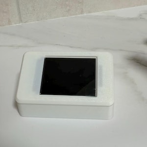 Op de afbeelding: Een witte rechthoekige doos met een zwart scherm. Het scherm is gecentreerd op de bovenkant van de doos. De doos staat op een wit oppervlak.