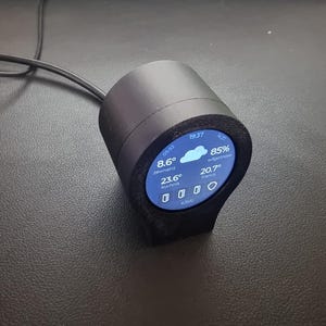 May include: A black, cylindrical smart device with a blue screen displaying weather information. The screen shows the time, temperature in Fahrenheit, humidity, and other weather details. A black cable extends from the device.