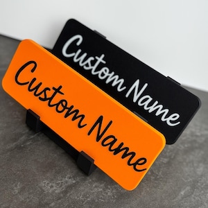 May include: Two rectangular nameplates, one orange and one black, each with the words "Custom Name" in a cursive font. The orange nameplate is in the foreground, and the black one is behind it. Both are held by black stands.