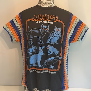 May include: A black t-shirt featuring a graphic print of a cat, owl, mouse, rabbit, frog, and raven with the text ADOPT A FAMILIAR and Black Magic Animal Rescue. The sleeves and side panels are accented with multi-coloured blue, orange, and grey crochet.