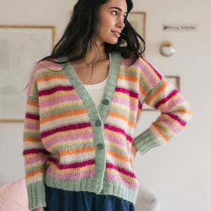 May include: A multi-coloured striped cardigan in shades of pink, orange, green, and white. The sweater has a V-neck, button closure, and long sleeves. The model is wearing the cardigan with a white top and blue denim shorts.