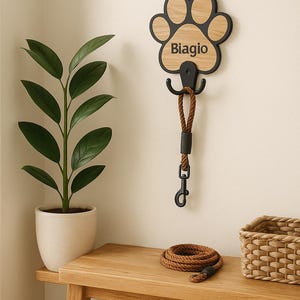 May include: A wooden paw-shaped leash holder with the name "Biagio" in black lettering. It has a black hook and a brown rope leash attached. A rolled-up brown leash and a woven basket sit on a wooden bench, with a potted plant to the left.