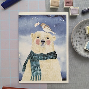 Può includere: Stampa artistica ad acquerello di un orso polare con una sciarpa blu, con un uccello appollaiato sulla sua testa che tiene una piccola bandiera. L'orso ha guance rosee e uno sfondo neutro. La stampa artistica è su una carta bianca.