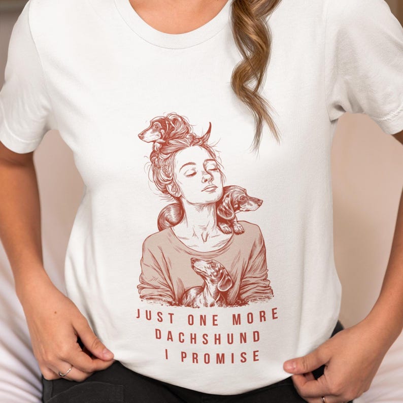 以下が含まれることがあります： 3匹のダックスフンドと女性の茶色のイラストが描かれた白いTシャツ。「JUST ONE MORE DACHSHUND I PROMISE」の文字が茶色でプリントされています。