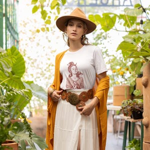 以下が含まれることがあります： グラフィックイラストが描かれた白いTシャツ、茶色の帽子、マスタード色のカーディガンを着た女性。また、円形の装飾が施された幅広の茶色のベルトと白いスカートを着用しています。背景には緑豊かな植物が写っています。
