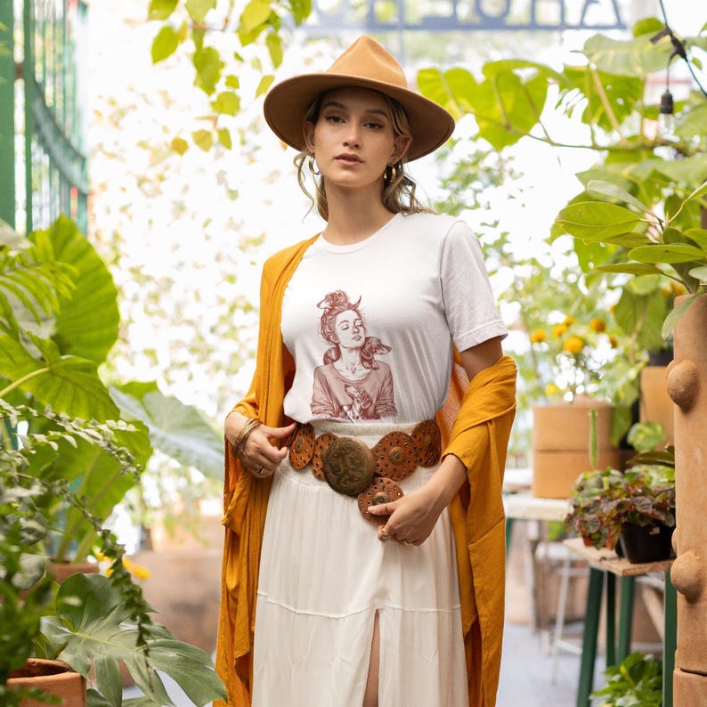 以下が含まれることがあります： グラフィックイラストが描かれた白いTシャツ、茶色の帽子、マスタード色のカーディガンを着た女性。また、円形の装飾が施された幅広の茶色のベルトと白いスカートを着用しています。背景には緑豊かな植物が写っています。