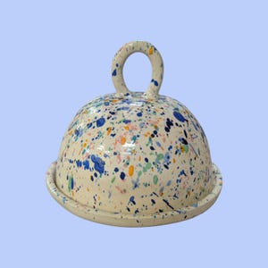 May include: A ceramic cloche with a cream-coloured base and dome, decorated with colourful paint splatters in blue, pink, green, and orange. The dome has a circular handle. The cloche sits on a matching base.