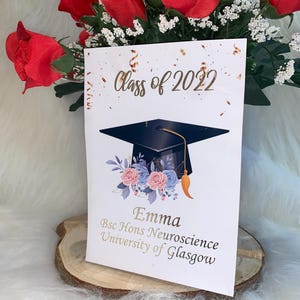 Könnte beinhalten: Eine Abschlusskarte mit dem Text "Class of 2022" und einer Illustration einer Abschlusskappe. Die Karte zeigt den Namen "Emma" und ihre Abschlussinformationen. Rote Rosen und weiße Blumen sind im Hintergrund.
