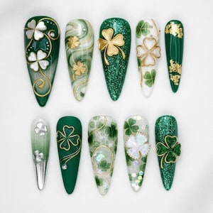 Könnte beinhalten: Eine Sammlung von zehn langen, spitz zulaufenden Kunstnägeln mit verschiedenen Designs zum St. Patrick's Day. Die Nägel weisen grüne, weiße und goldene Farben mit Kleeblatt- und Glitzerakzenten auf. Einige Nägel haben Goldfolien- oder Edelsteinverzierungen.