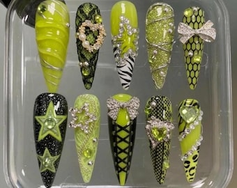 Op de afbeelding: Een set van tien lange, amandelvormige kunstnagels in een doorzichtige plastic doos. De nagels zijn voornamelijk neon groen, zwart en zilver, met verschillende ontwerpen, waaronder glitter, kant, strikken en ster- en hartaccenten.
