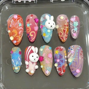 Puede incluir: Un juego de diez uñas postizas en forma de almendra con diseños coloridos. Cada uña presenta obras de arte únicas, incluyendo patrones florales, estrellas, corazones y un conejito 3D. Las uñas están en una caja de plástico transparente.