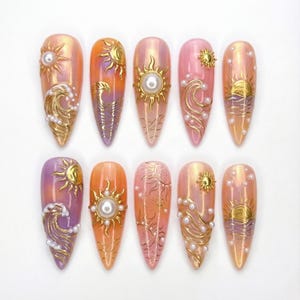 Puede incluir: Un conjunto de diez uñas artificiales largas en forma de almendra. Cada uña presenta un diseño de puesta de sol con soles dorados, olas y detalles de perlas. La paleta de colores incluye tonos de naranja, rosa y morado, creando un aspecto vibrante y artístico.
