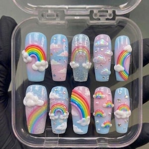 Könnte beinhalten: Ein Set aus zehn künstlichen Nägeln mit einem Himmelsdesign. Jeder Nagel zeigt einen Regenbogen, Wolken und Sterne auf hellblauem oder rosa Hintergrund. Einige Nägel haben Glitzerakzente, in einer transparenten Kunststoffbox.