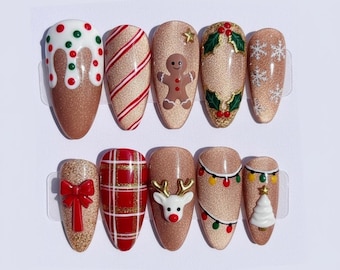 May include: A set of ten artificial nails with Christmas-themed designs. The nails feature glittery gold bases with various festive decorations, including a gingerbread man, snowflakes, and a red bow. Other designs include candy cane stripes and a plaid pattern.
