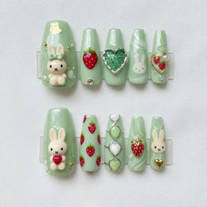 May include: A set of ten pale green press-on nails with diverse designs. Some nails feature 3D bunny and strawberry accents, while others have glitter hearts and swirl patterns. The nails are arranged on a clear plastic sheet.