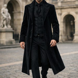 May include: A black velvet overcoat with embroidered detailing on the collar and cuffs. The coat is worn over a black vest, shirt, and tie. The outfit is completed with black trousers and shoes. The setting is outdoors, with a stone walkway and buildings in the background.