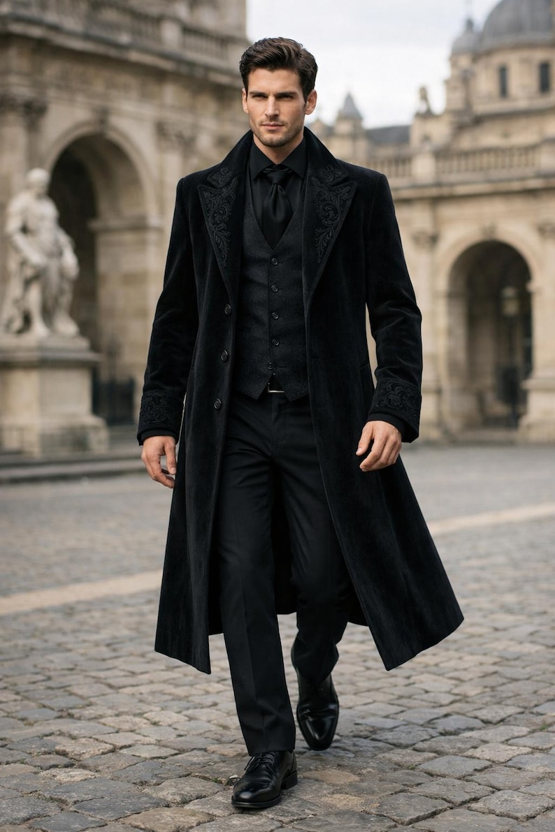 May include: A black velvet overcoat with embroidered detailing on the collar and cuffs. The coat is worn over a black vest, shirt, and tie. The outfit is completed with black trousers and shoes. The setting is outdoors, with a stone walkway and buildings in the background.
