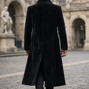 May include: A long, black, velvet coat with a classic design. The coat features a notched collar, long sleeves, and a split back. The person is walking on a cobblestone street in front of a building with arches and statues.
