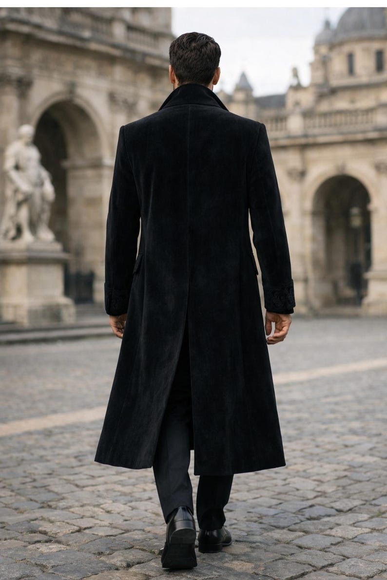 May include: A long, black, velvet coat with a classic design. The coat features a notched collar, long sleeves, and a split back. The person is walking on a cobblestone street in front of a building with arches and statues.