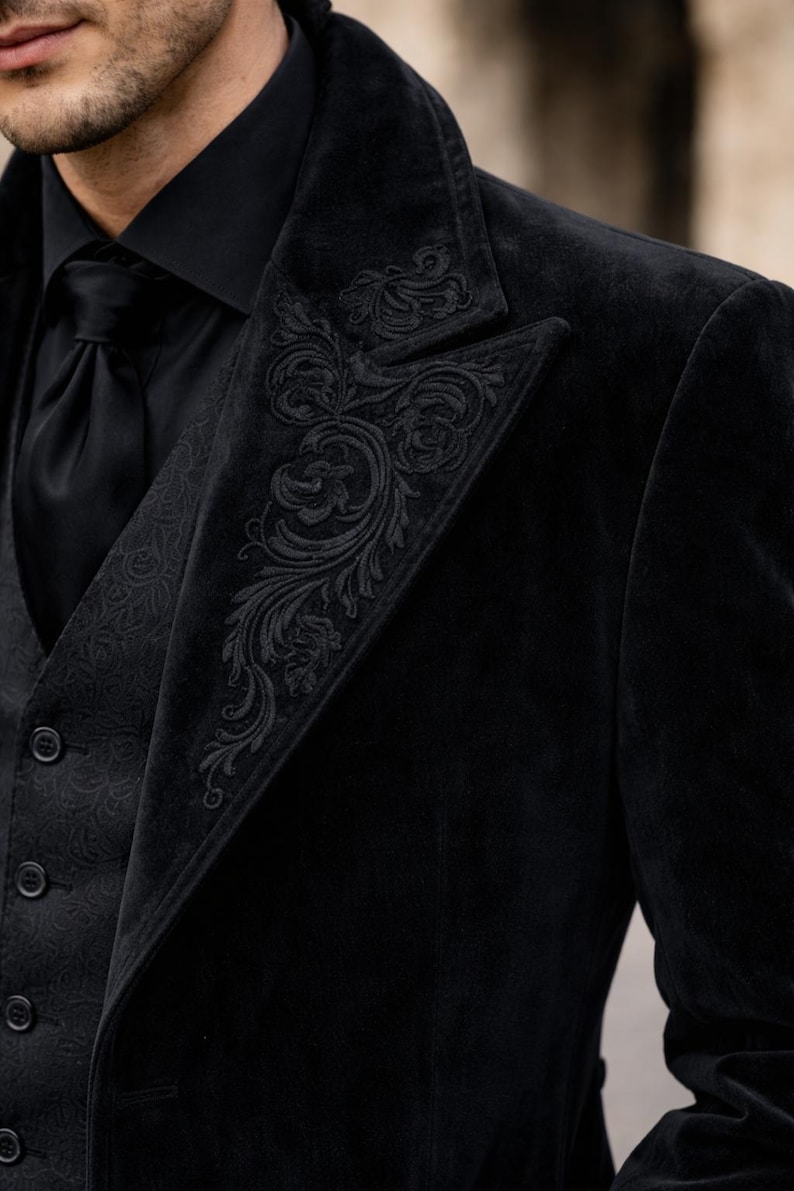 May include: Black velvet suit with ornate embroidered detailing on the lapel and vest. The suit includes a black tie and a black collared shirt. The suit is a formal style.