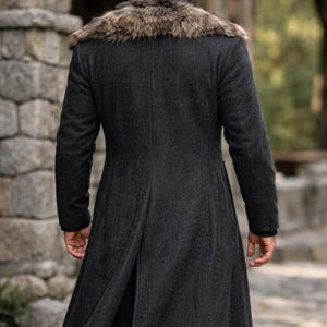 May include: A dark gray wool coat with a faux fur collar. The long coat has a tailored fit and a split back. The person is facing away from the camera, and the background is a stone wall and greenery.
