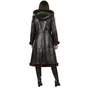 May include: A long, black, crocodile-textured coat with a hood and brown fur trim. The coat is knee-length and has a flared skirt. The model is wearing black boots. The coat is made of a leather-like material.