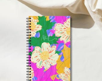 May include: A spiral-bound notebook featuring a vibrant floral pattern with large white flowers, green leaves, and abstract shapes in bright pink, yellow, and blue. The word Notes is written in a thin white cursive script across the center.