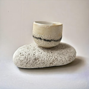 Può includere: Una piccola tazza in ceramica strutturata con un design beige e nero, appoggiata su una pietra porosa grigio chiaro. La tazza ha una forma arrotondata e una fascia scura vicino alla base. La pietra ha una superficie ruvida e puntinata.