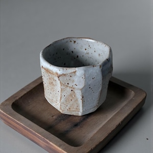 Può includere: Una piccola tazza in ceramica bianco sporco con un design geometrico e una texture maculata è appoggiata su un vassoio quadrato di legno. La tazza ha una forma sfaccettata unica e un aspetto rustico fatto a mano. Il vassoio è di colore marrone caldo.