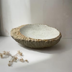 Pode incluir: Uma tigela de cerâmica redonda e rasa com um exterior esculpido e texturizado em tom bege claro. A superfície interior é lisa, de cor branco sujo, e salpicada com pequenos pontos castanhos escuros. O rebordo é espesso e irregular, feito à mão.