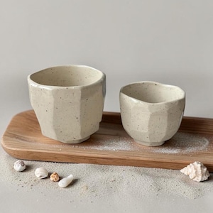 Op de afbeelding: Twee gebroken witte keramische bekers met een gespikkelde afwerking en gefacetteerde, onregelmatige zijkanten. De bekers hebben een brede, open rand en een taps toelopende voet, wat zorgt voor een ambachtelijke uitstraling.