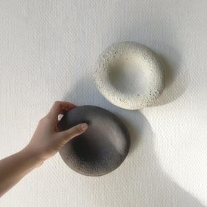 May include: Two circular, bowl-shaped objects with a textured, porous surface. One object is off-white and the other is dark charcoal gray. Both items feature a smooth, concave center and a rough, uneven outer rim, presenting a distinct contrast.