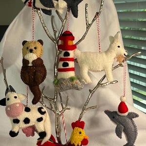 May include: A collection of handmade felt ornaments, including a shark, bear, lighthouse, goat, cow, rooster, and dolphin, displayed on a silver decorative tree. Each ornament is detailed with various colors and textures, and suspended by red and white twine.