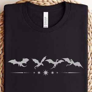 Könnte beinhalten: Schwarzes T-Shirt mit Rundhalsausschnitt und silberner Grafik von fünf fliegenden Drachen über einer gepunkteten Linie mit Sternen. Das T-Shirt ist auf einer gewebten, kreisförmigen Oberfläche gefaltet.