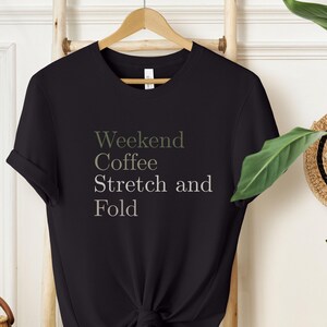 Könnte beinhalten: Schwarzes T-Shirt mit den Worten "Weekend Coffee Stretch and Fold" in hellgrauer Schrift. Das Shirt hängt an einem Holzbügel vor einer weißen Wand. Weidenkörbe und eine Strohtasche sind im Hintergrund.