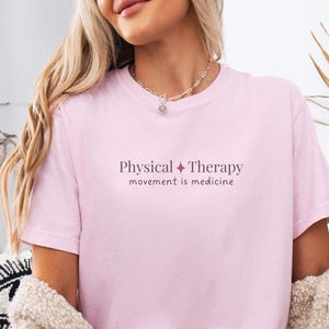 May include: A light pink t-shirt with the words "Physical Therapy" and "movement is medicine" printed in a dark pink font. The shirt is short-sleeved and made of a soft material.