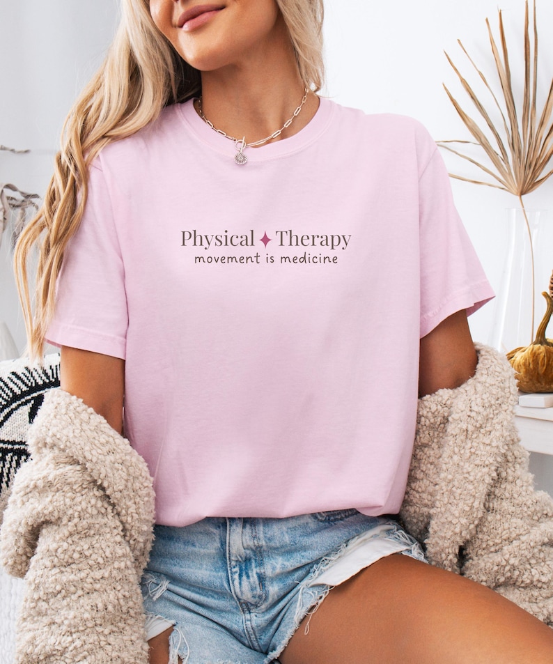 May include: A light pink t-shirt with the words "Physical Therapy" and "movement is medicine" printed in a dark pink font. The shirt is short-sleeved and made of a soft material.