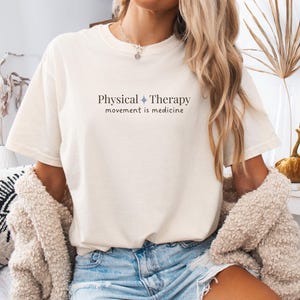 May include: Cream-colored t-shirt with the text "Physical Therapy movement is medicine" in dark gray. The shirt is paired with denim shorts. The overall aesthetic is casual and comfortable.