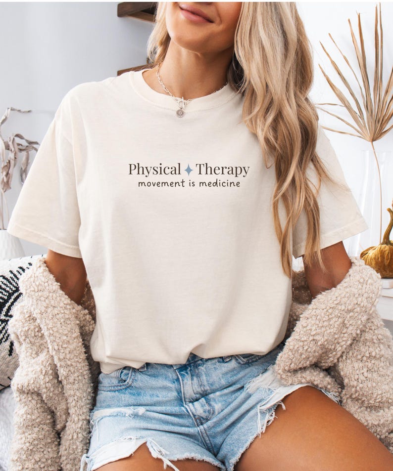 May include: Cream-colored t-shirt with the text "Physical Therapy movement is medicine" in dark gray. The shirt is paired with denim shorts. The overall aesthetic is casual and comfortable.