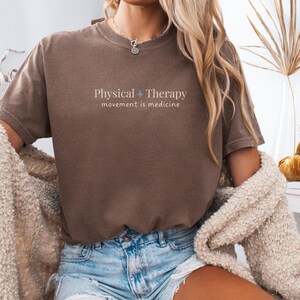 May include: Brown t-shirt with the text "Physical Therapy movement is medicine" in a light color. The shirt is paired with denim shorts. A beige cardigan is draped over the shoulders.