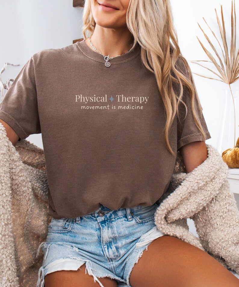 May include: Brown t-shirt with the text "Physical Therapy movement is medicine" in a light color. The shirt is paired with denim shorts. A beige cardigan is draped over the shoulders.