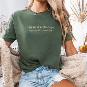 May include: Olive green t-shirt with the text "Physical Therapy" and "movement is medicine" in a white, minimalist font. The shirt is paired with denim shorts. A beige cardigan is draped over the shoulders.
