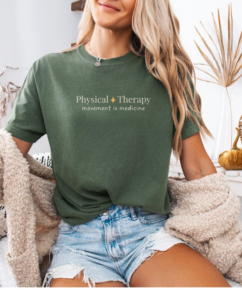 May include: Olive green t-shirt with the text "Physical Therapy" and "movement is medicine" in a white, minimalist font. The shirt is paired with denim shorts. A beige cardigan is draped over the shoulders.