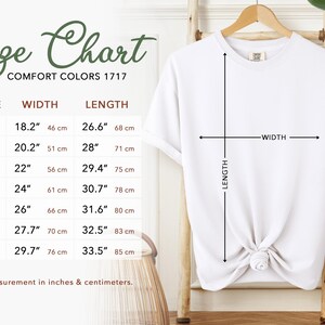 May include: White t-shirt hanging on a wooden hanger, with a size chart to the left. The size chart shows measurements in inches and centimeters for sizes S to 4XL. The shirt's width and length are indicated with arrows.