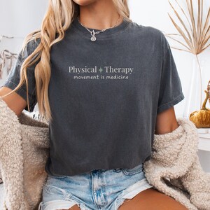 May include: Dark gray t-shirt with the words "Physical Therapy" and "movement is medicine" printed in light green. The shirt has a relaxed fit and short sleeves. The model is wearing denim shorts and a cream-colored cardigan.