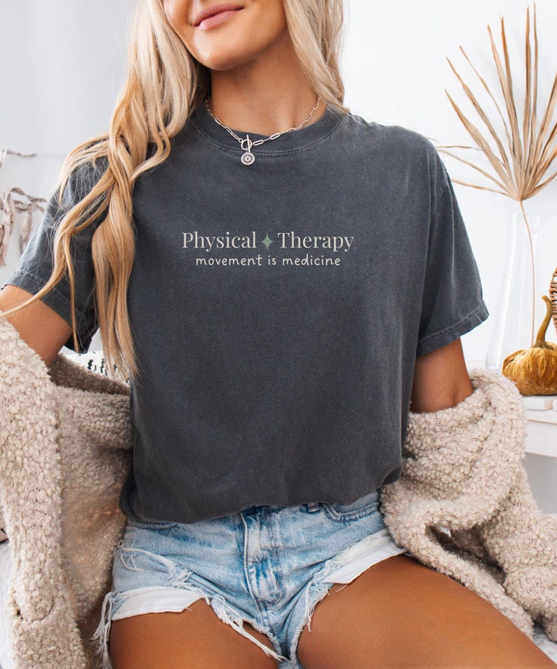 May include: Dark gray t-shirt with the words "Physical Therapy" and "movement is medicine" printed in light green. The shirt has a relaxed fit and short sleeves. The model is wearing denim shorts and a cream-colored cardigan.